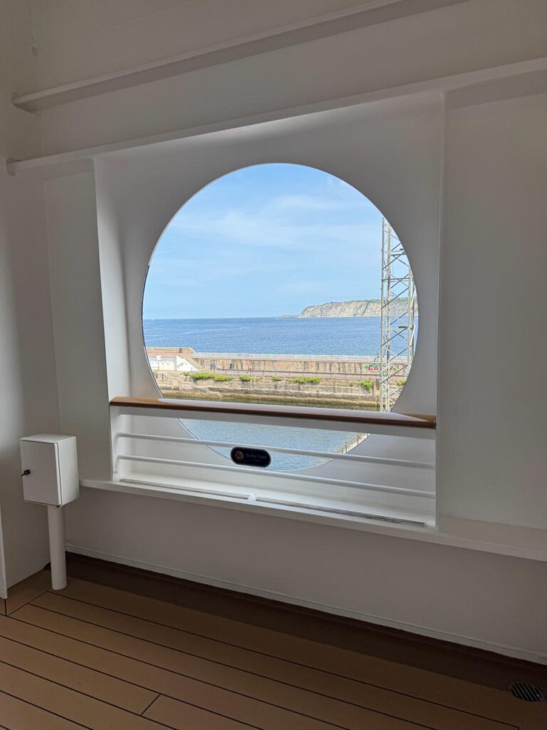 Open circular porthole on the Disney Dream cruise ship’s Deck 4, showing an unobstructed view of the ocean and distant shoreline through the wide opening framed by white walls and a wooden railing, with no glass barrier visible.
