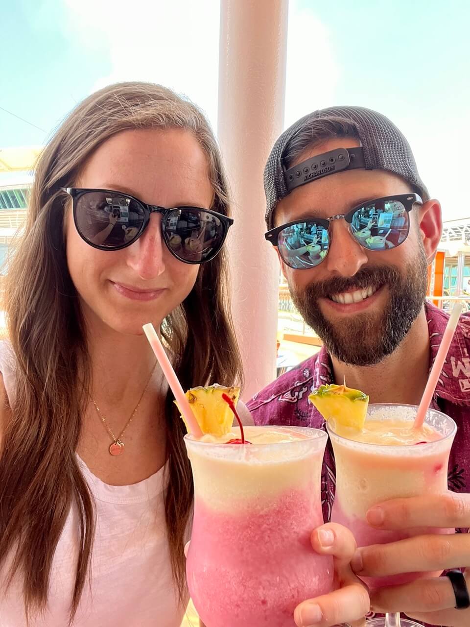 Two adults wearing sunglasses on a cruise ship deck, smiling and holding frozen tropical drinks garnished with pineapple.