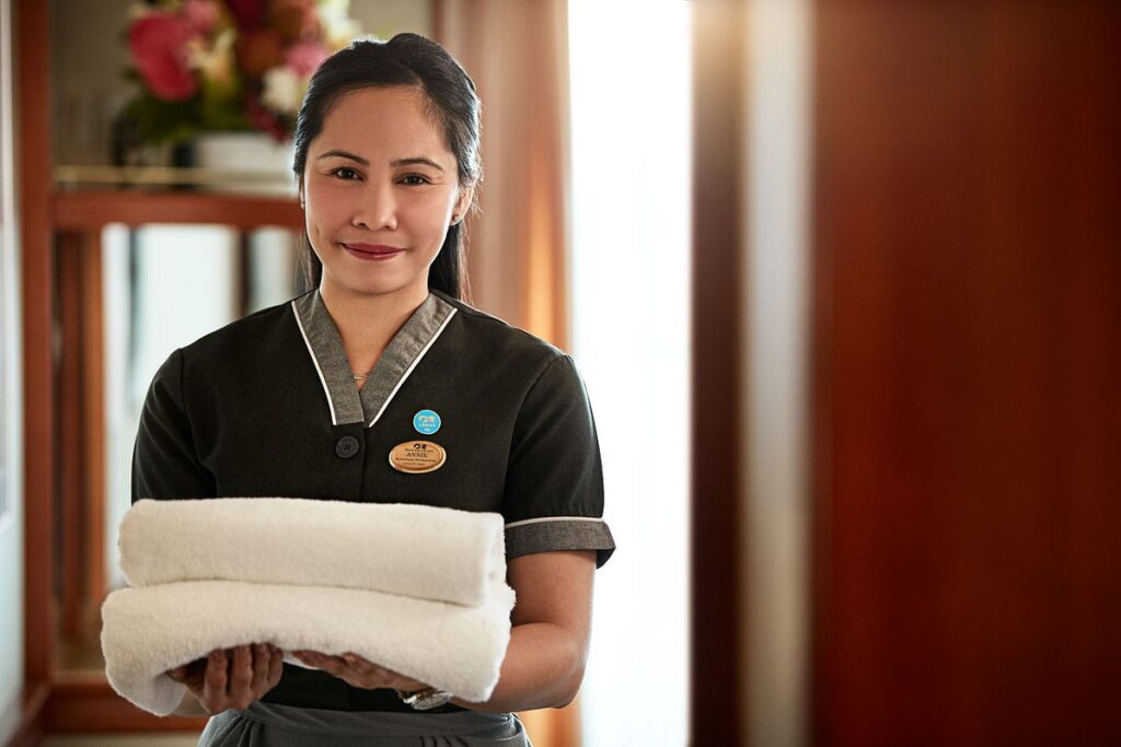 Princess cruise ship cabin steward stands in a stateroom doorway holding two folded white towels.