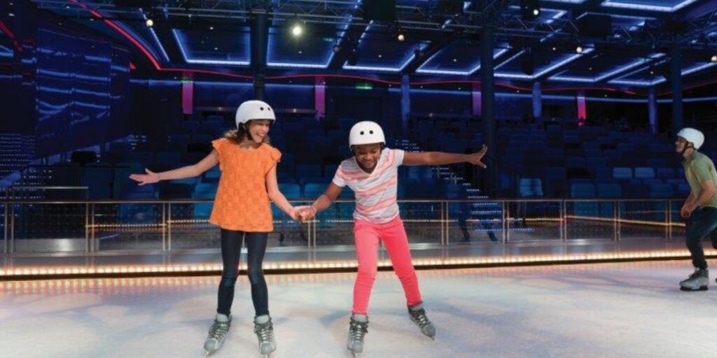 Two kids wearing helmets ice skate hand-in-hand on an indoor rink aboard a Royal Caribbean cruise ship, laughing and having fun under vibrant neon lighting. A man skates nearby, with empty stadium seating in the background, capturing a playful moment at sea.