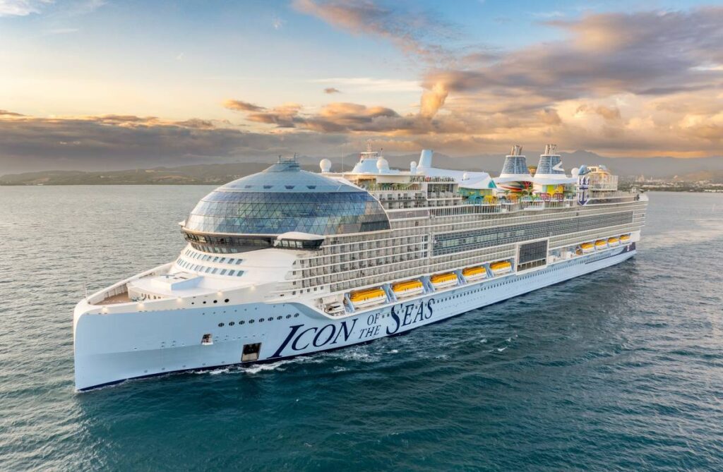 Royal Caribbean's Icon of the Seas cruise ship sails gracefully at dusk, with the glow of the setting sun casting a warm light on its sleek, white structure. The ship's impressive size and modern design, featuring a unique glass dome, are set against a backdrop of distant mountains and a pastel sky.