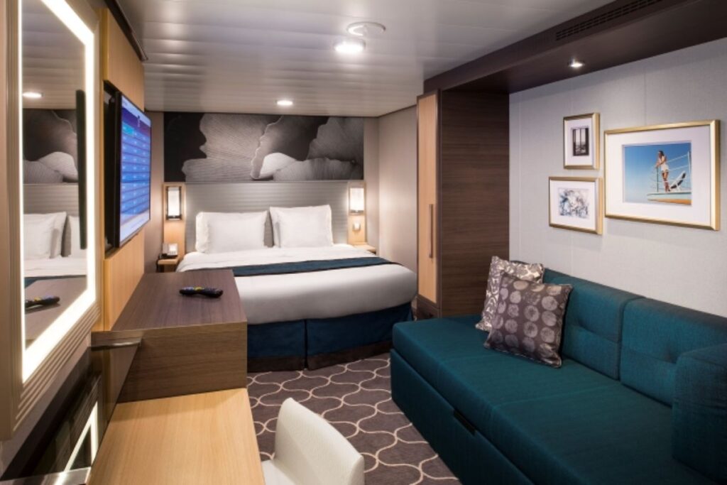 This image shows an interior stateroom aboard the Harmony of the Seas cruise ship. The room features a cozy queen-sized bed with crisp white linens, flanked by bedside tables and lamps. There's also a small seating area, a wall-mounted flat-screen TV, and modern decor that creates a comfortable and inviting atmosphere for guests.