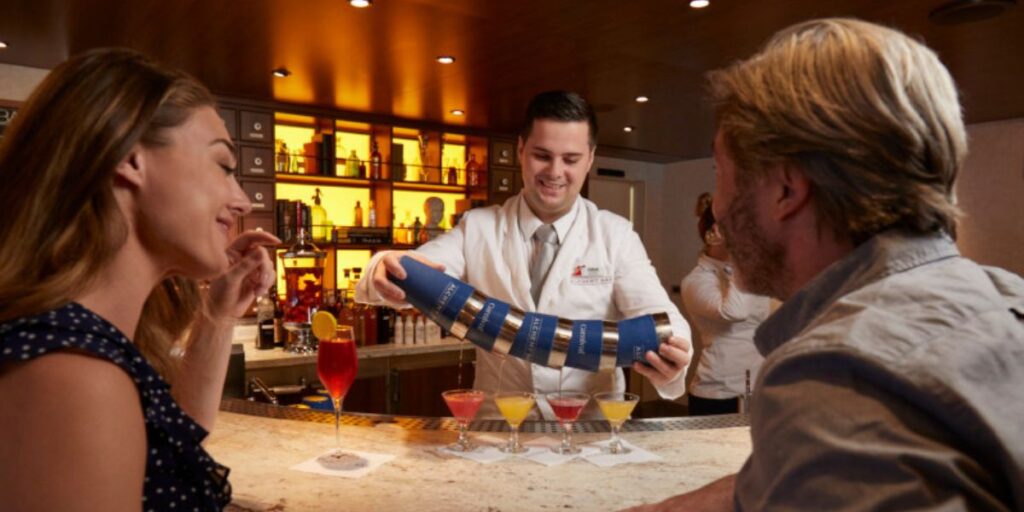 A bartender in a white coat demonstrates a cocktail-flipping trick to an amused woman and man seated at a warmly lit bar, with a selection of colorful cocktails displayed on the countertop.