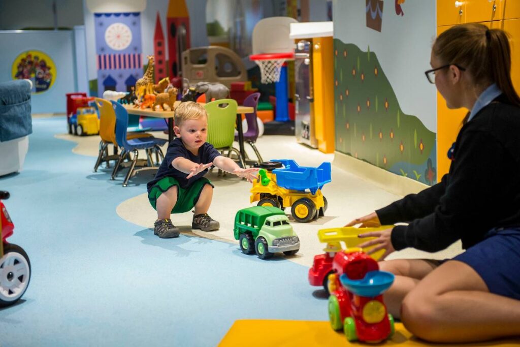 A toddler joyfully playing with colorful toy trucks in the 'It's a Small World Nursery' on Disney Magic, with a crew member nearby, in a vibrant room filled with whimsical Disney-themed decorations.