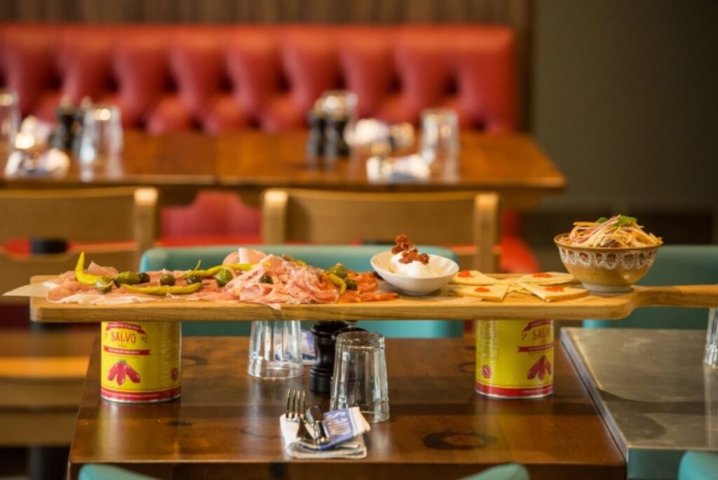An inviting charcuterie board with a selection of cured meats, olives, and cheese, presented on a wooden plank supported by canned tomatoes, set on a table at Jamie's Italian restaurant onboard Harmony of the Seas, with a red tufted booth in the background.