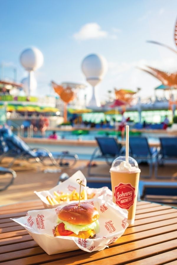 A classic Johnny Rockets meal—complete with a cheeseburger, fries, and a milkshake—sits on a wooden table beside a Royal Caribbean pool deck. The sunny cruise atmosphere features lounge chairs, colorful umbrellas, and modern ship architecture in the background.
