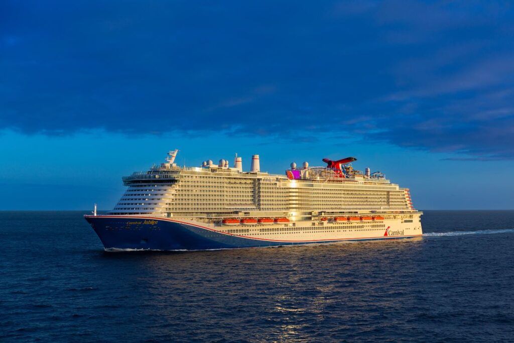 The Carnival Jubilee cruise ship is captured sailing across the ocean under a deep blue sky during what appears to be the golden hour, as the ship is bathed in warm light. The ship’s iconic red funnel and vibrant exterior are prominently displayed, with several decks visible, each lined with lifeboats. The scene highlights the grandeur of the ship against the expansive and tranquil sea.