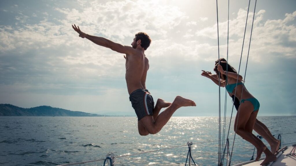 A man in swim trunks leaps off a boat into the water, arms outstretched and legs bent mid-air, while a woman in a bikini playfully pushes him. The sun casts a warm glow on the sea, creating a shimmering reflection. In the background, a coastline with rolling hills is visible under a partly cloudy sky, capturing the essence of a carefree summer adventure.
