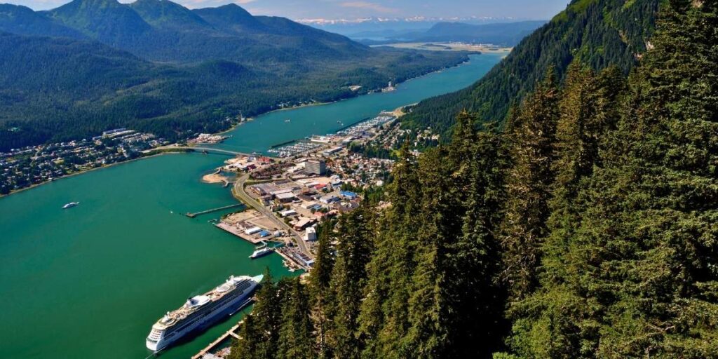 This image shows a sweeping aerial view of Juneau, Alaska, where lush evergreen forests frame the city nestled between the Gastineau Channel and dramatic mountain peaks. A cruise ship is docked at the harbor, emphasizing Juneau’s role as a key stop for Alaskan cruises.