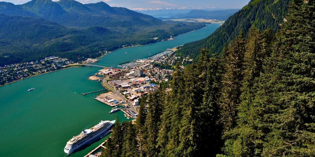 This image shows a sweeping aerial view of Juneau, Alaska, where lush evergreen forests frame the city nestled between the Gastineau Channel and dramatic mountain peaks. A cruise ship is docked at the harbor, emphasizing Juneau’s role as a key stop for Alaskan cruises.