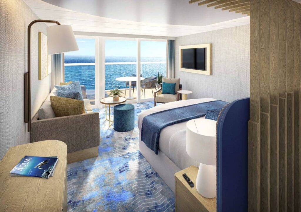 A Junior Suite on Icon of the Seas offers a cozy and stylish setting, with a comfortable bed adorned in blue, a sitting area with ocean-inspired decor, and large windows that frame the stunning sea view, inviting natural light into the room.