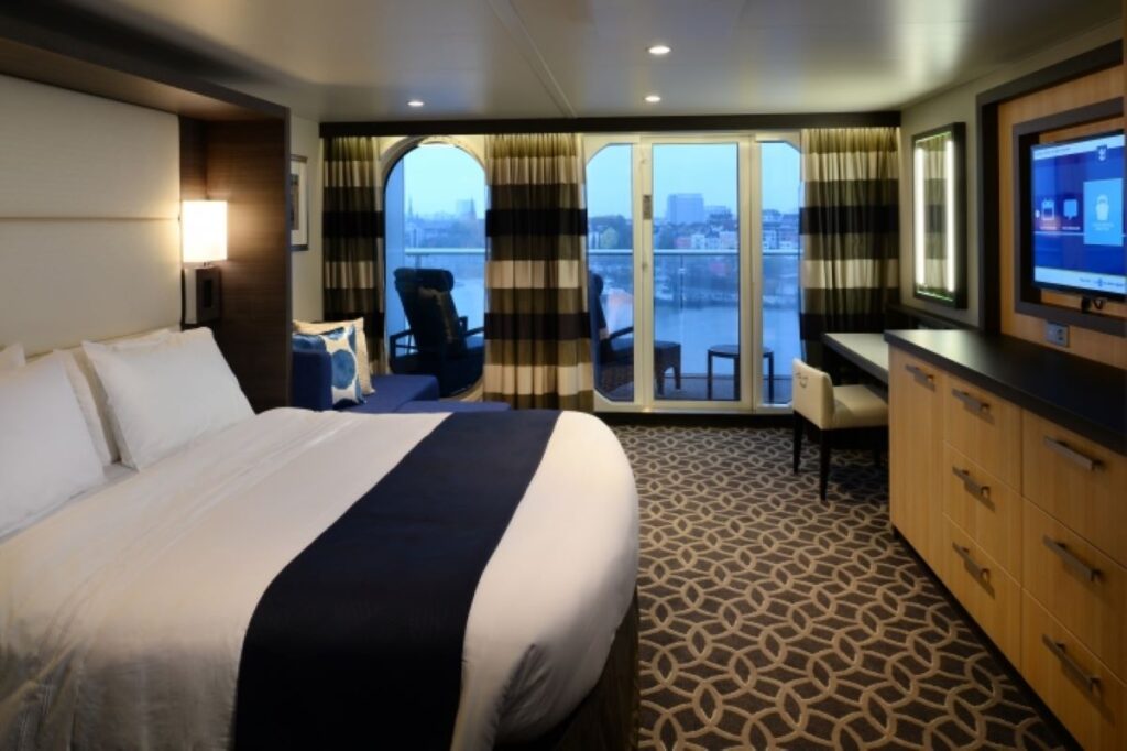 Elegant Junior Suite on Quantum of the Seas cruise ship, with a plush king-sized bed, patterned carpet, a cozy sitting area by the balcony door, and a cityscape view through the round window.