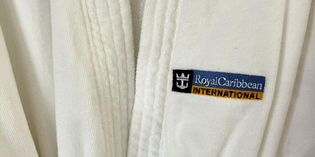 Close-up of a plush white bathrobe with the Royal Caribbean International logo embroidered on a patch. The logo features a stylized crown and anchor symbol above the brand name, indicating the premium amenities provided aboard their cruise ships.