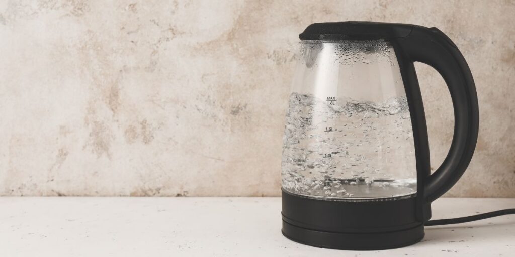 Glass electric kettle with black accents shown mid-boil, with bubbling water visible through the transparent sides. The kettle is placed against a light stone-textured wall on a white countertop.
