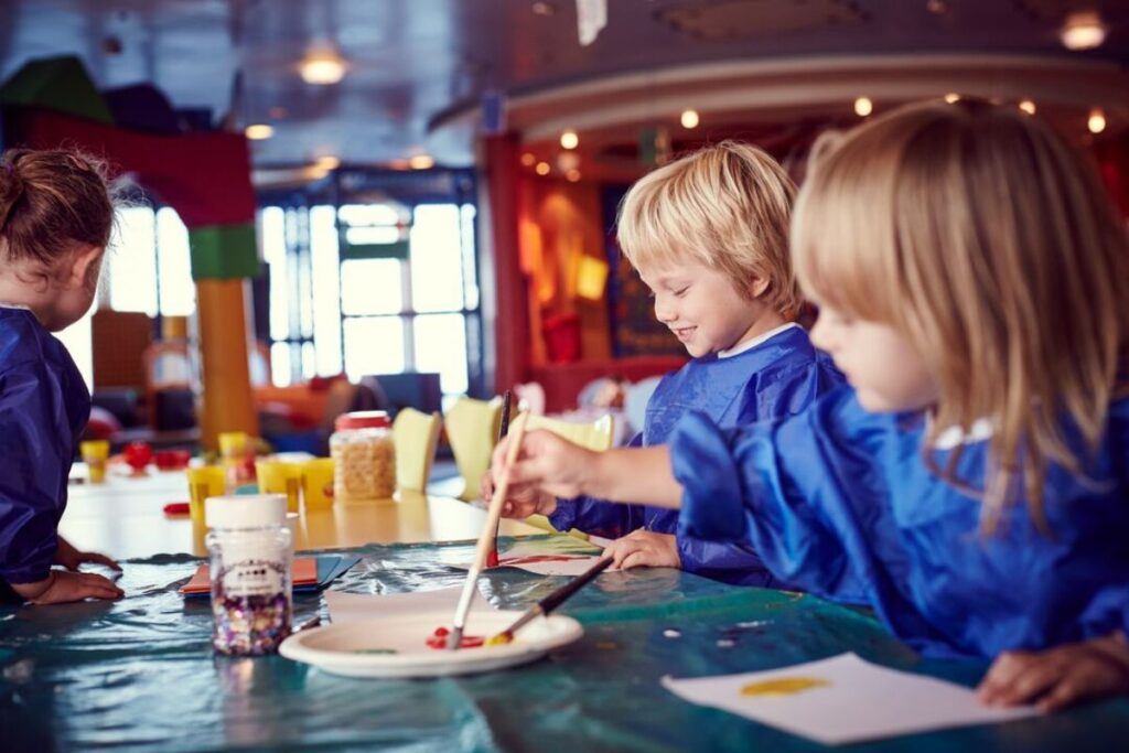 The image shows children engaged in creative activities at a kid's club on a P&O cruise ship. The kids, dressed in blue smocks, are painting and playing with colorful materials on a table covered with craft supplies. The vibrant and cheerful setting includes bright furniture and playful decor, designed to provide a fun and engaging environment for children onboard.