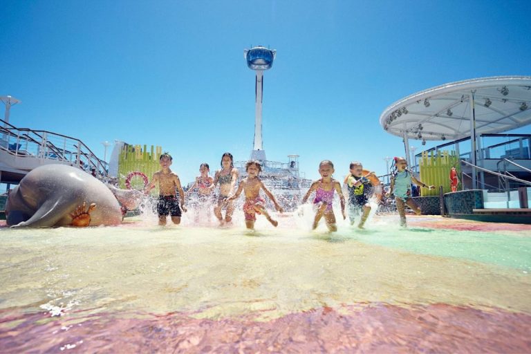 A group of children joyfully running through a shallow water play area on a Royal Caribbean cruise ship, with colorful water slides and attractions in the background under a clear blue sky.