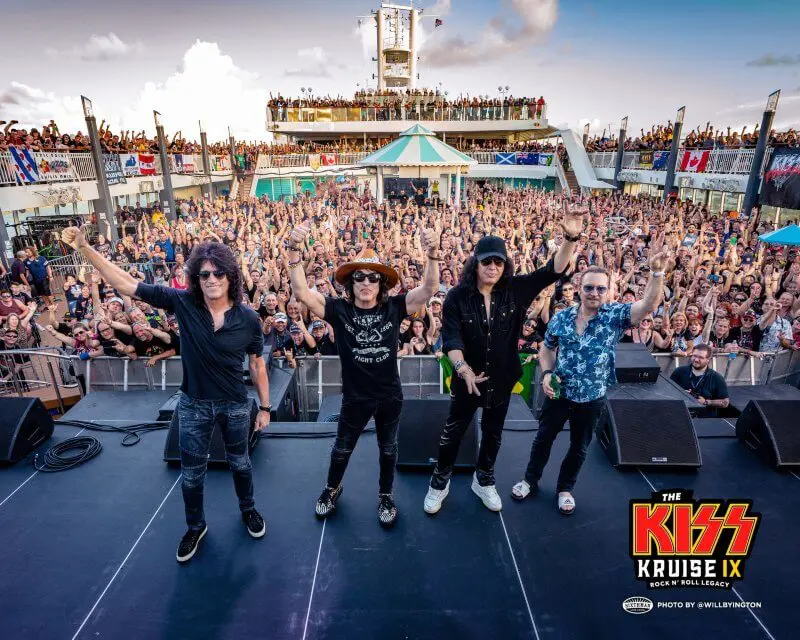 Four members of the rock band KISS stand on an outdoor stage aboard a cruise ship, raising their arms to a massive crowd of fans cheering behind them. The deck is packed with people, some waving flags, as the ship sails under a partly cloudy sky. The bottom right corner features the "KISS Kruise IX" logo, indicating this is part of the band's themed cruise experience.