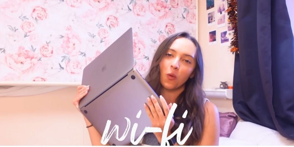 A woman sitting in her cruise ship cabin holding an open laptop, with the word “Wi-Fi” displayed across the image. She appears to be talking while explaining something, with floral wallpaper and photos behind her.