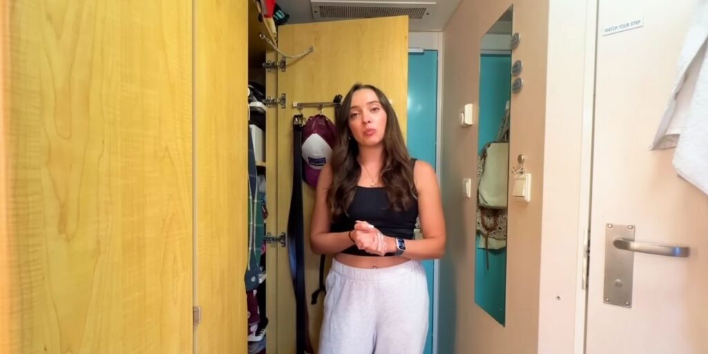 A woman standing inside a compact cruise ship cabin, wearing a black tank top and light sweatpants. She faces the camera while speaking, with an open closet and neatly stored belongings visible behind her.