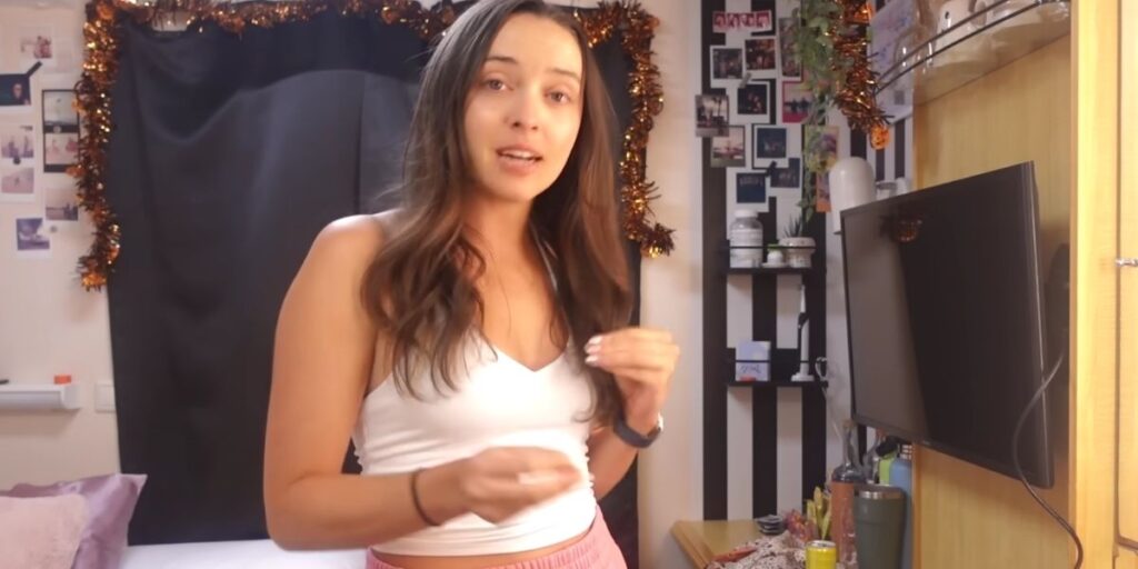 A woman standing inside a cozy, decorated cruise ship cabin, wearing a white tank top and pink shorts. She faces the camera while speaking, with photos, plants, and festive garland on the wall behind her.
