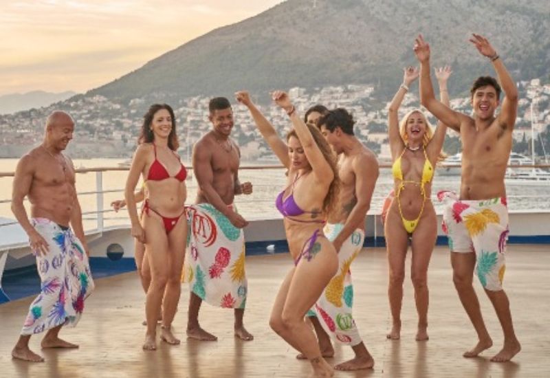 A group of people in vibrant swimwear and tropical-print sarongs dance and celebrate on the open deck of an **LLV Club Cruise**. The scenic backdrop features mountains and a coastal town, enhancing the exotic and luxurious atmosphere. With smiles, raised hands, and energetic movements, the group radiates a carefree, party vibe in a sensual, adults-only setting.