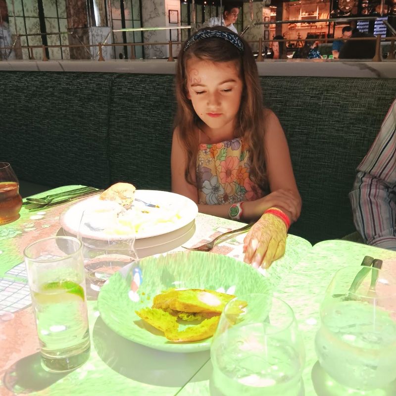 A girl sits at a dining table, focused on her meal at Le Petit Chef on the Celebrity Apex cruise ship. The table features animated projections, adding a playful and immersive element to her dining experience, while glasses of water and a partially eaten dish are visible in front of her.