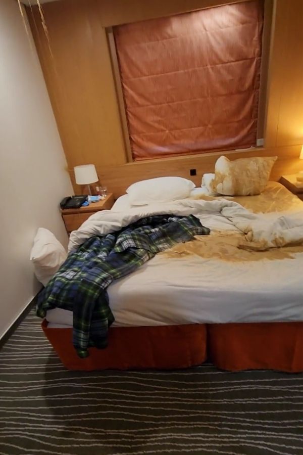 A stateroom on the Marella Explorer 2 cruise ship shows extensive water damage, with a soaked bed, stained bedding, and visible ceiling leaks dripping onto the mattress. A plaid shirt lies on the bed, emphasizing the disruption caused by the leak aboard the Marella Explorer 2 cruise ship.