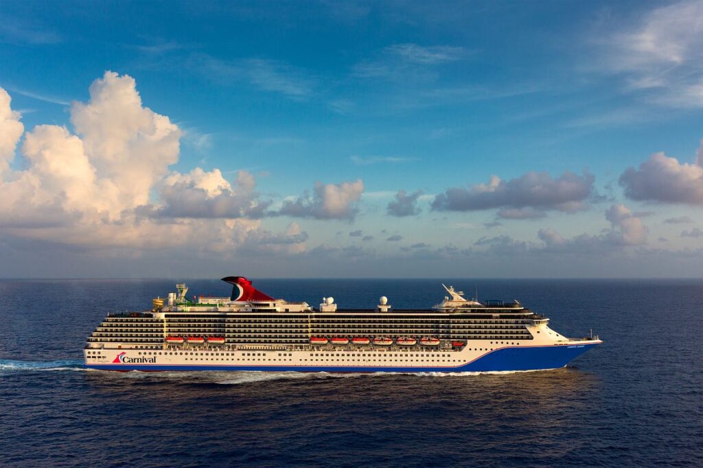 The Carnival Legend cruise ship sails under a vast sky, fluffy clouds overhead, reflecting the golden sunlight on its journey across the endless blue ocean.