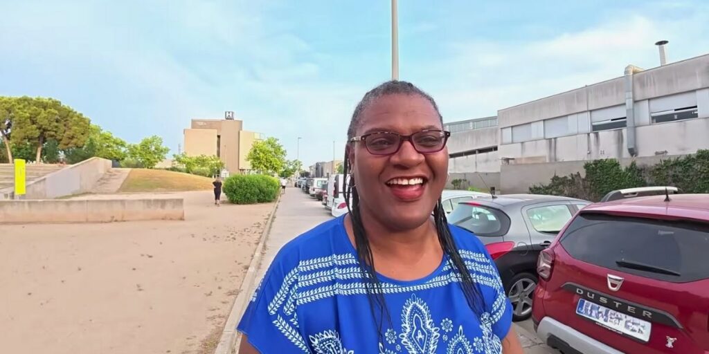 A smiling woman wearing glasses and a blue patterned blouse stands outdoors beside a row of parked cars, with modern buildings and a tree-lined path in the background. The scene is bright and sunny, suggesting a casual, cheerful moment.