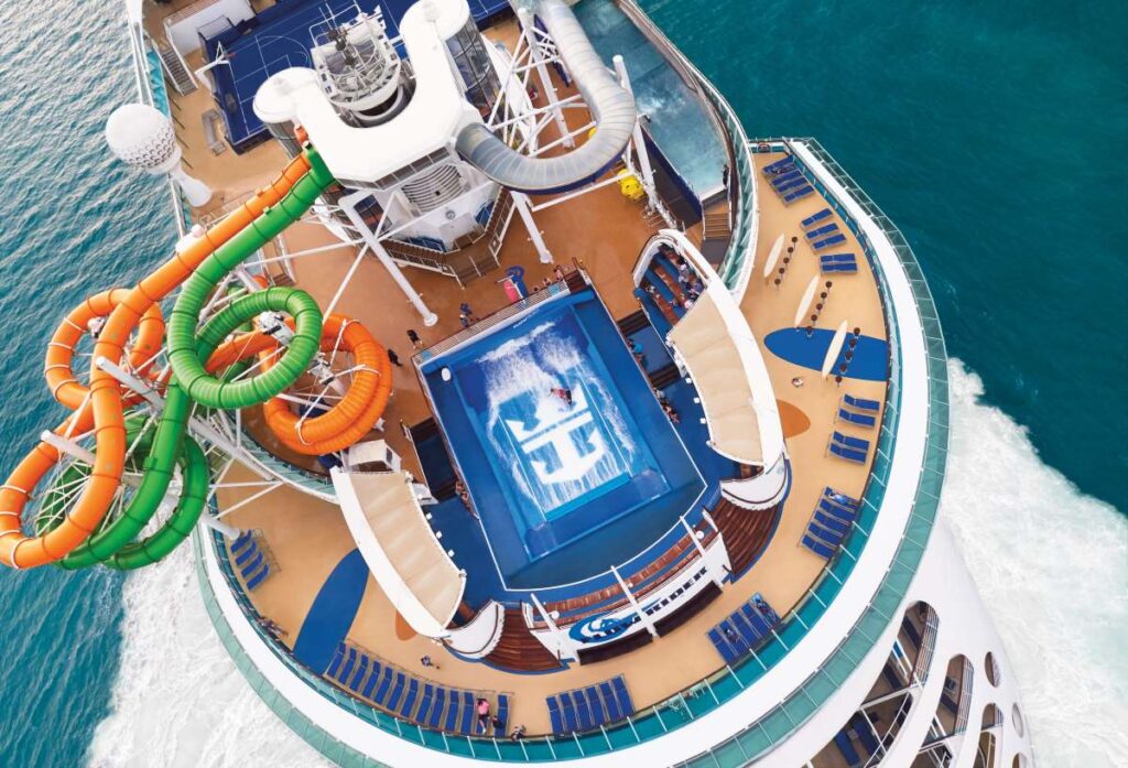 An aerial view of the deck on Royal Caribbean's Liberty of the Seas, highlighting an array of twisting water slides in vibrant orange and green. Below, passengers enjoy the ship's spacious pool area, with the ocean's expanse providing a captivating backdrop to the bustling onboard activities.