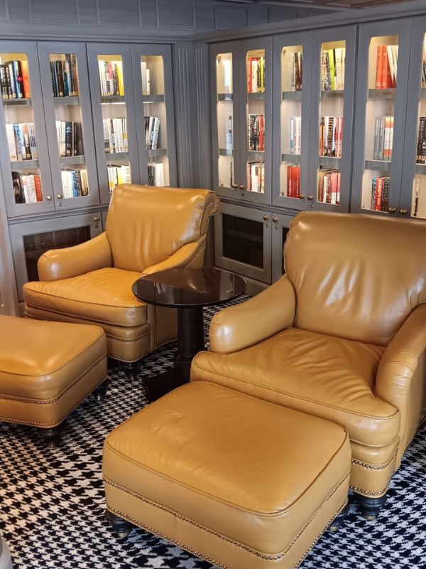 This image shows a cozy library on the Oceania Mariner cruise ship. It features two large, comfortable tan leather armchairs with matching ottomans, positioned on a black-and-white houndstooth-patterned carpet. The chairs are situated next to a small round table, and behind them are bookshelves filled with a variety of books, providing an inviting atmosphere for reading and relaxation.