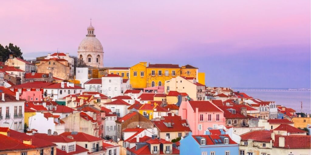 Colorful buildings with red rooftops in Lisbon, Portugal, at sunset with the dome of the National Pantheon in the background.