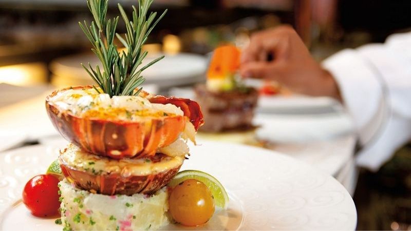 A gourmet lobster tail dish is elegantly plated atop seasoned mashed potatoes and garnished with a sprig of rosemary, cherry tomatoes, and a lime wedge, as a chef adds final touches in the background. The upscale presentation suggests a fine dining experience, likely aboard a cruise or in a high-end restaurant.