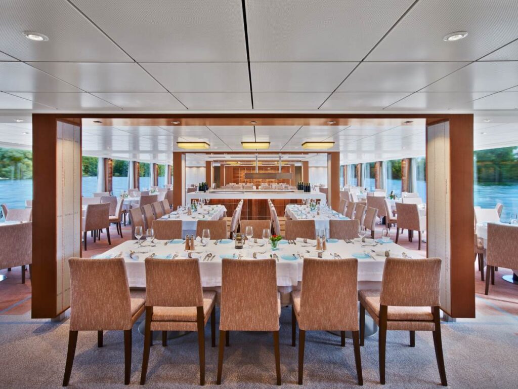 This image shows The Restaurant aboard the Viking Kadlin, featuring a modern and bright dining space with neatly set tables. The room has floor-to-ceiling windows that provide natural light and scenic views, paired with minimalist décor and warm tones. The tables are dressed in crisp white linens, accented with polished glassware and small floral arrangements for a refined yet inviting dining ambiance.