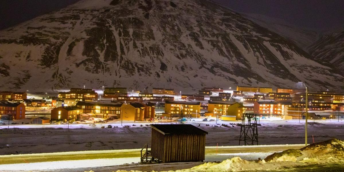 Nighttime view of Longyearbyen, Svalbard, with warmly lit buildings nestled at the base of a snow-covered mountain. The town glows under a dark Arctic sky, with snowmobiles and a small wooden shed visible in the snowy foreground.