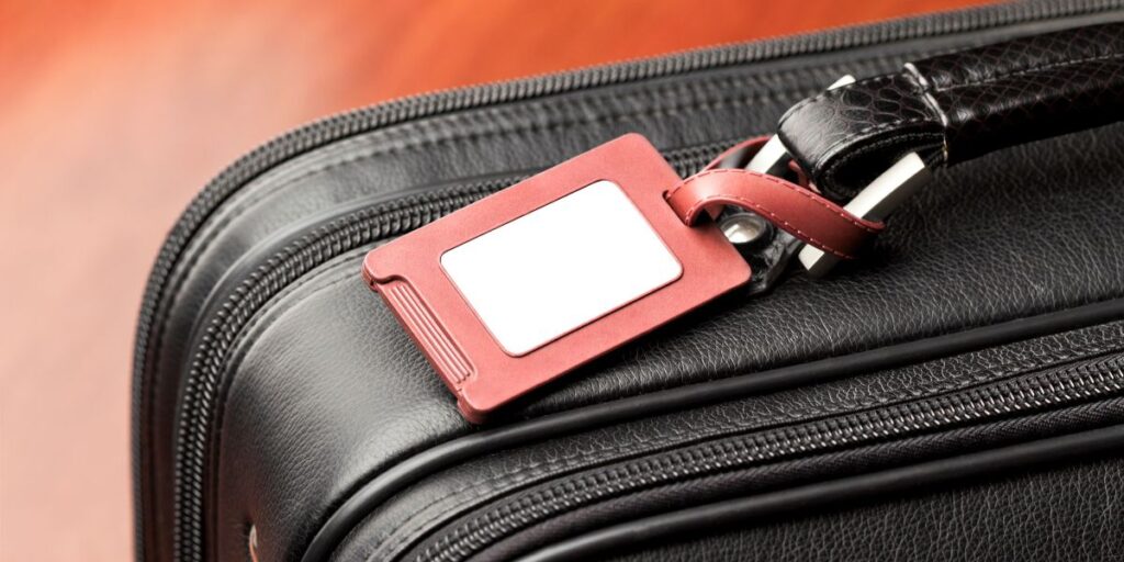 Close-up of a black textured suitcase with a red leather luggage tag attached, featuring a blank white label for personal information against an orange background.