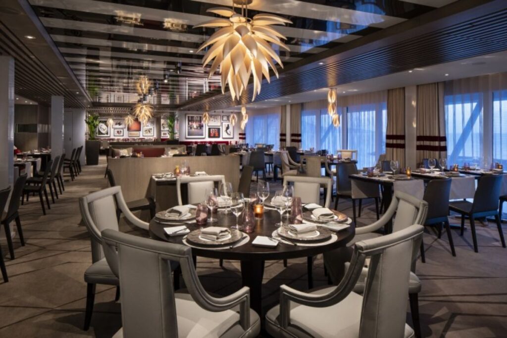 This image features the elegant dining room of Luminae at The Retreat on Celebrity Edge. The restaurant showcases sophisticated decor with plush seating, beautifully set tables, and ambient lighting from unique, leaf-shaped chandeliers. The room's warm, modern atmosphere is enhanced by floor-to-ceiling windows, providing a stylish and intimate dining experience for guests.