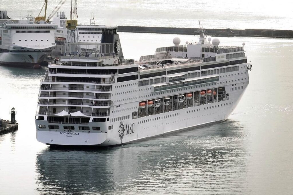 The MSC Armonia cruise ship gracefully navigates a harbor, showcasing its gleaming white structure against a backdrop of smaller vessels and maritime equipment. The ship, marked prominently with the MSC logo, features multiple decks lined with rows of windows, enhancing its elegant design as it moves through the calm sea waters near the port.