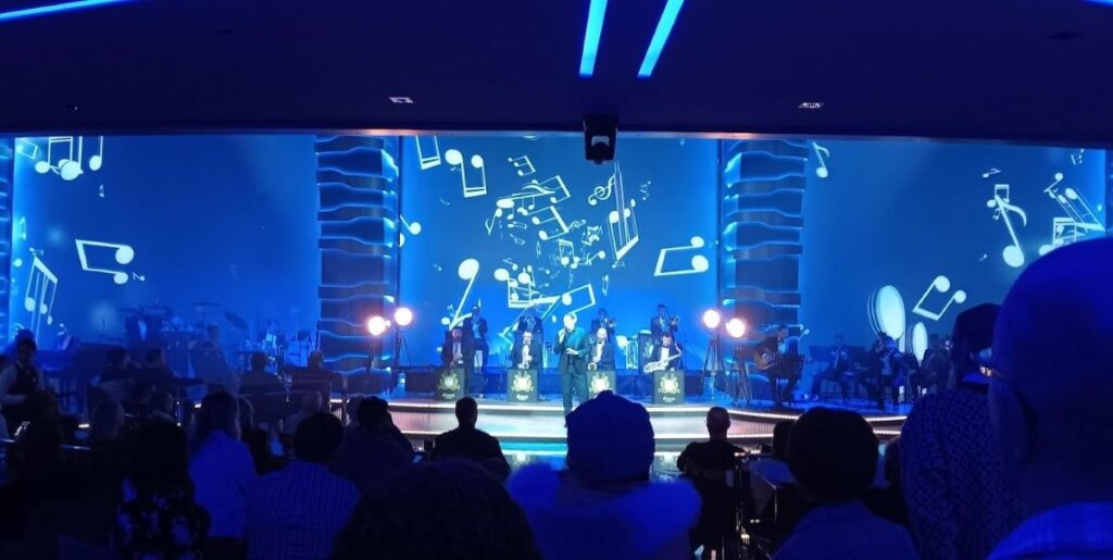 Guests enjoy a live performance in the Carousel Lounge on an MSC cruise ship, with the stage bathed in blue light and adorned with dynamic musical note decorations, as silhouetted spectators watch from the dimly lit audience area.