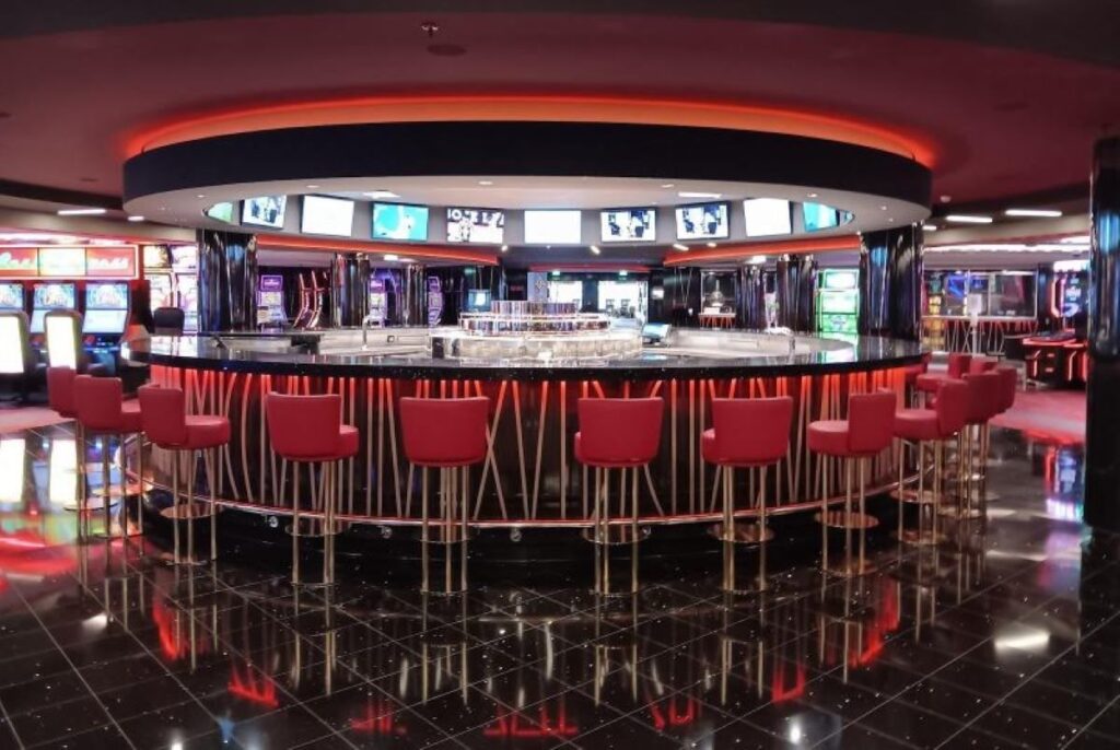 A vibrant casino bar aboard an MSC cruise ship, featuring a circular bar with glossy red stools and a reflective black floor that mirrors the colorful neon lights and slot machines in the background.