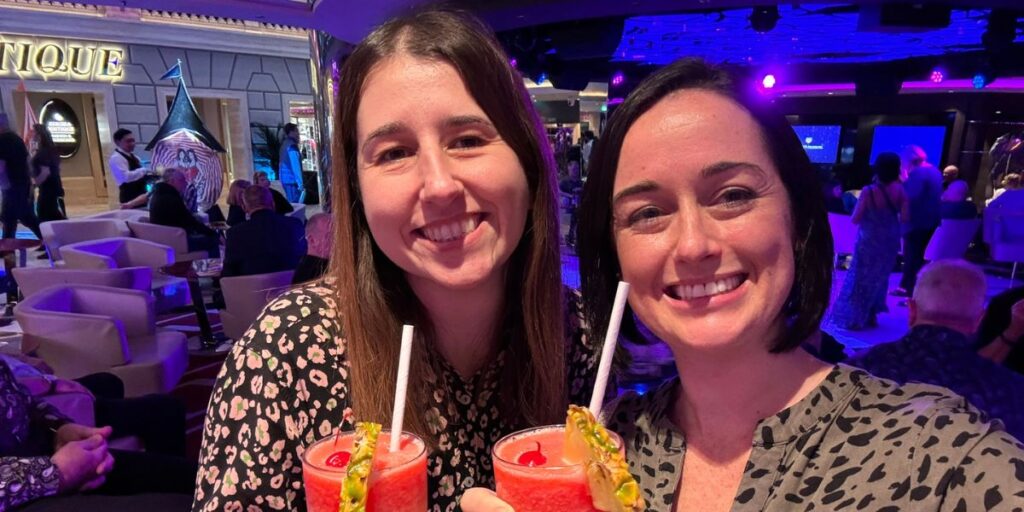 Two smiling women holding tropical drinks with straws, garnished with fruit, in a lively and colorful bar area onboard a cruise ship, with other guests and a distinctive spiral sculpture in the background.