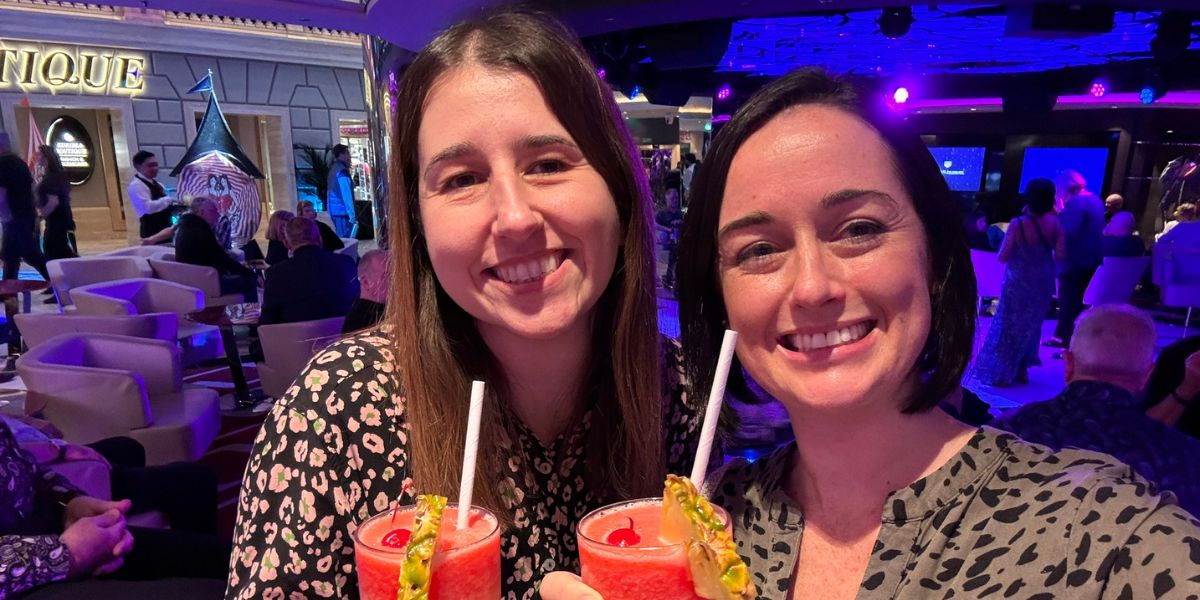 Two smiling women holding tropical drinks with straws, garnished with fruit, in a lively and colorful bar area onboard a cruise ship, with other guests and a distinctive spiral sculpture in the background.