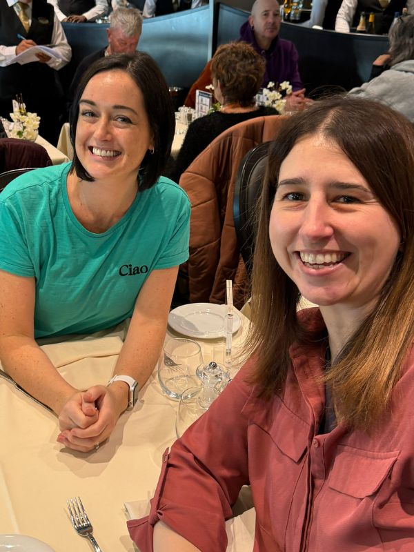 Two smiling women seated at a dining table in a busy restaurant, one wearing a teal 'Ciao' t-shirt and the other in a pink shirt. They appear to be enjoying a social event, with tables set with white linens and glassware surrounding them.