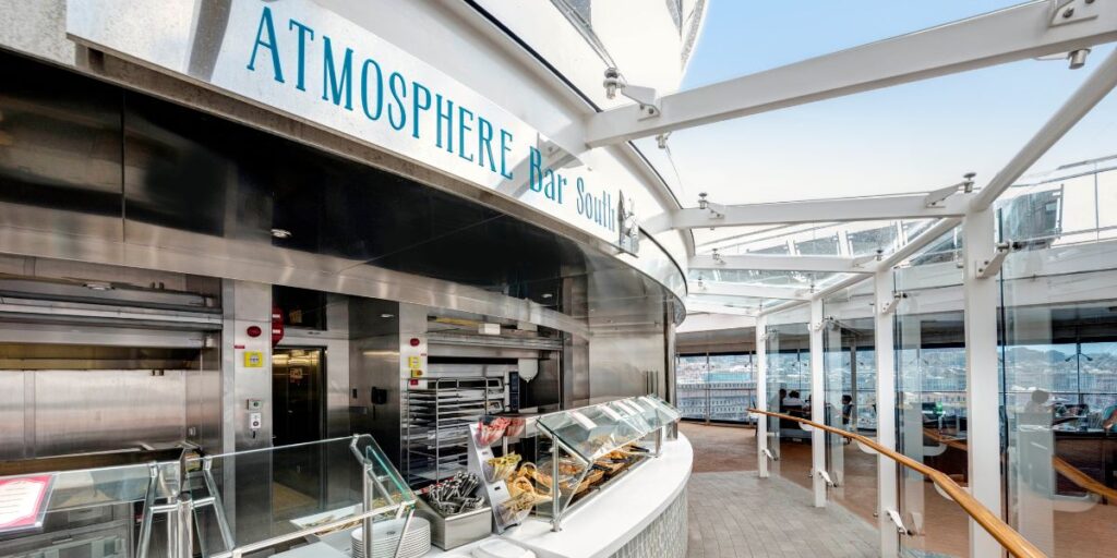 Interior view of the Atmosphere Bar South on MSC Euribia, showcasing a clean and unoccupied food serving area with a selection of pastries on display, flanked by stacked white plates and catering utensils, with guests seated in the background behind glass partitions.