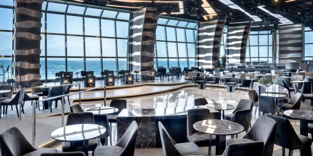 The sophisticated Carousel Lounge on MSC Euribia, with its expansive ocean view through floor-to-ceiling windows, features a modern design with a circular central bar, dark upholstered chairs, and marble tables, all under soft ambient lighting.