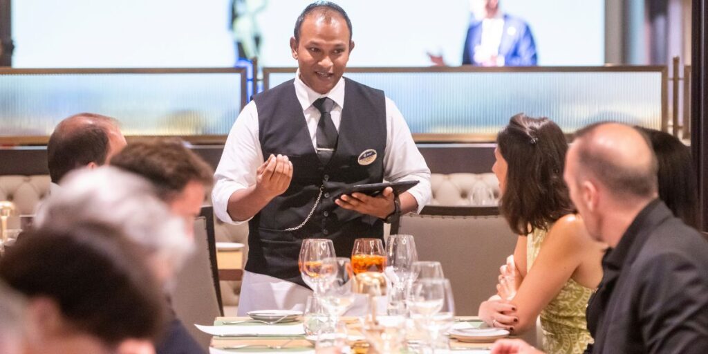 A waiter engaging with guests at 'Le Grill' restaurant, holding a digital tablet for order taking. The dining table is set elegantly with wine glasses and cutlery, while patrons appear to be enjoying a meal.