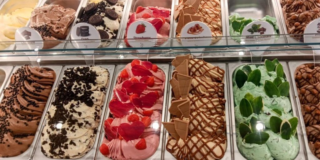 A selection of gelato flavors displayed in a case, with options including rich chocolate adorned with cookie pieces, fresh strawberry topped with fruit slices, a drizzled caramel variant and more.