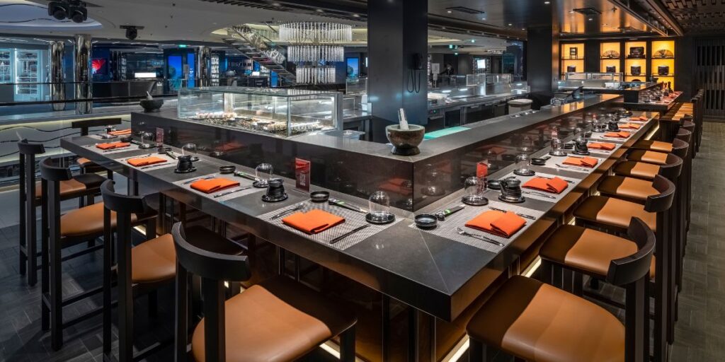 A modern sushi bar setup with a sleek black counter, high stools with tan seats, and vibrant orange napkins neatly laid out. The bar showcases an open kitchen with a well-lit display of fresh ingredients, offering a contemporary dining experience.