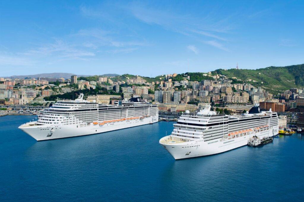 Aerial view of two MSC cruise ships, MSC Fantasia and MSC Poesia, docked side by side in a port against the scenic backdrop of a densely built hillside city. The cruise ships, towering with multiple decks, are impressive with their pristine white exteriors and large scale, contrasting with the urban landscape of the city's colorful buildings and green hills in the background.