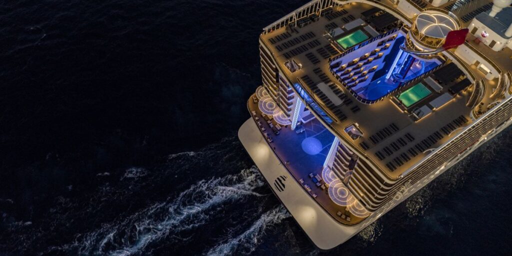 A stunning aerial night view of the aft section of an MSC cruise ship, illuminated by elegant blue and golden lights. The ship's multi-level decks, pools, and lounge areas create a luxurious ambiance as it sails through the dark ocean, leaving a gentle wake behind. The glow of the ship contrasts beautifully against the deep black water, highlighting its modern and sophisticated design.